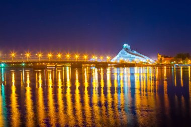 Parlak Daugava Nehri Köprüsü ve gece yansımalar. Letonya Riga'da modern Milli Kütüphane