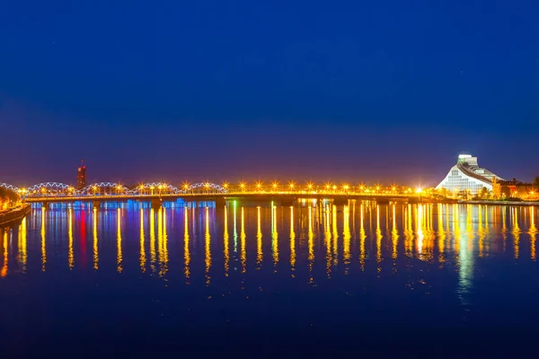 Ponte brilhante e reflexos noturnos no rio Daugava. Biblioteca Nacional Moderna da Letônia em Riga — Fotografia de Stock