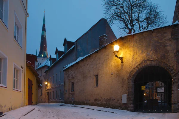 Old Tallinn étroite rue avec des lanternes brillantes et l'église Oleviste au premier plan. Soirée d'hiver enneigée . — Photo