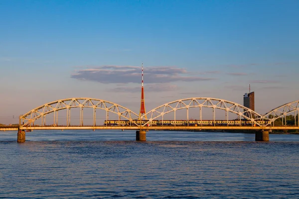 O comboio está a passar pela ponte sobre Daugava. Torre de TV e hora do pôr do sol. Riga, Letónia — Fotografia de Stock
