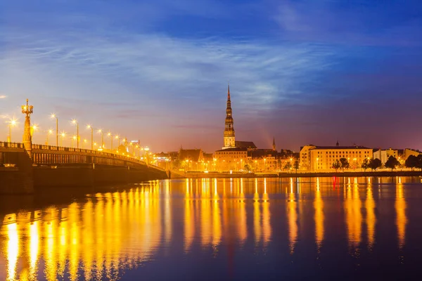 Büyük taş köprü ve St. Peter Kilisesi ile eski Riga gece manzarası. Işıklı şehir güneş battıktan sonra. Daugava Nehri üzerinden panoramik görünüm