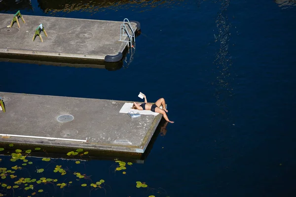 Young woman laying on pier, reading book, black swimsuit. Stockholm, Sweden. — Stock Photo, Image