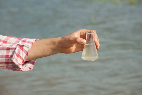 Prueba de pureza del agua. Frasco químico de mano con líquido, lago o río en el fondo . — Foto de Stock