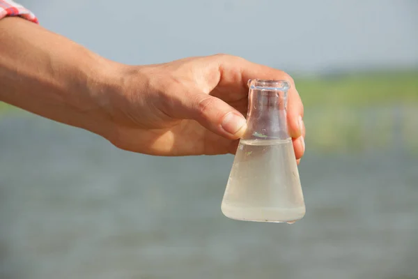 Vatten renhet Test. handen håller kemiska kolv med vätska, sjö eller älv i bakgrunden. — Stockfoto