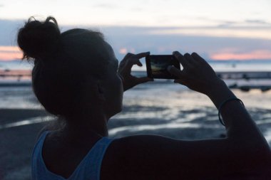 Kadın bir gün batımı bir telefon kamera denizden alır.