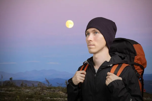 Młody człowiek z plecakiem w górach. Nocny pagórkowaty krajobraz z księżyca. — Zdjęcie stockowe