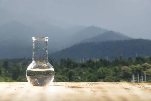 Acqua pulita in una fiaschetta di vetro da laboratorio su un tavolo di legno su sfondo montano. Concetto ecologico, test di purezza e qualità dell'acqua . — Foto Stock