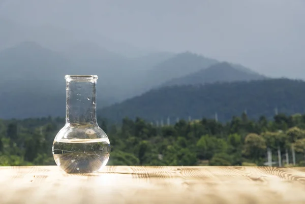 Clean water in a glass laboratory flask on wooden table on mountain background. Ecological concept, the test of purity and quality of water.