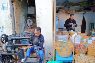 SFAX, TUNISIA - DECEMBER 25, 2019: A cobbler at work inside the medina with a sewing machine and stalls of nuts on the right side