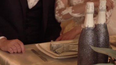 Masanın üzerinde bir buket ve iki bardak pembe şampanya var. Gelin ve damat arka planda oturuyor..