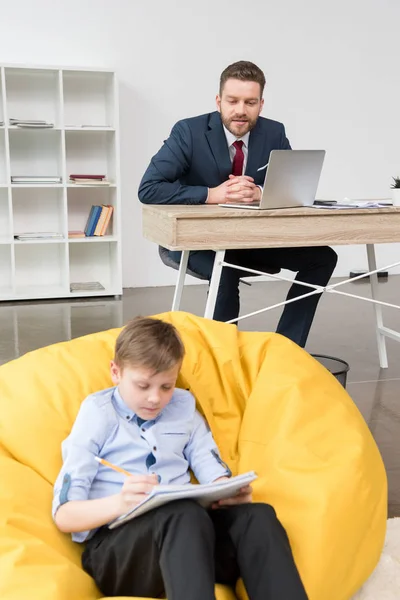 Boy drawing while his father working — Stock Photo, Image