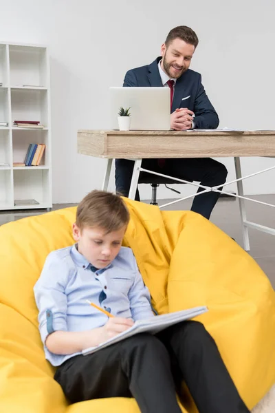 Niño dibujando mientras su padre trabaja - foto de stock