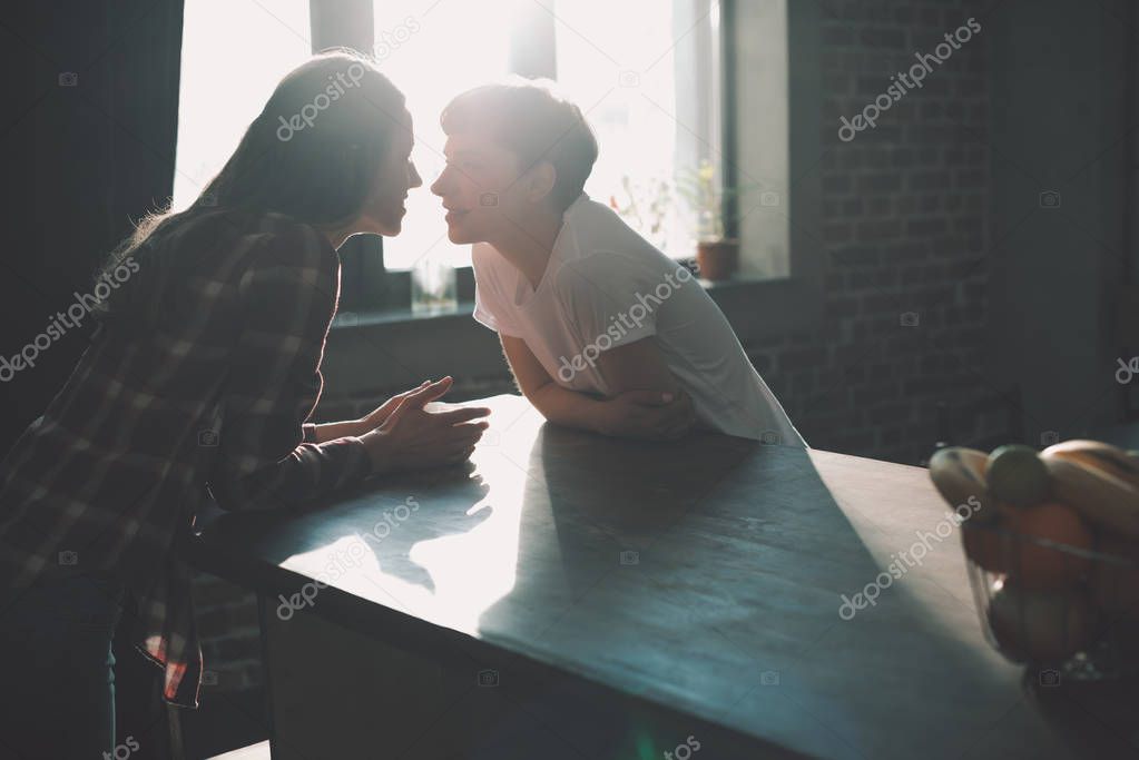 Silhouettes of young lesbian couple spending time together and kissing on kitchen in morning