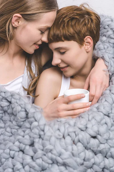 Крупный план счастливой лесбийской пары с чашкой кофе, обнимающей под вязаным шерстяным одеялом — стоковое фото