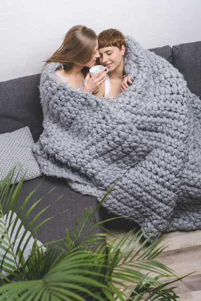 Glückliches lesbisches Paar mit einer Tasse Kaffee, das sich unter einer Strickdecke auf der Couch umarmt — Stockfoto