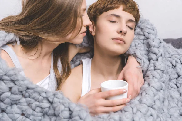 Giovane coppia lesbica con tazza di caffè che abbraccia sotto coperta di lana lavorata a maglia — Foto stock