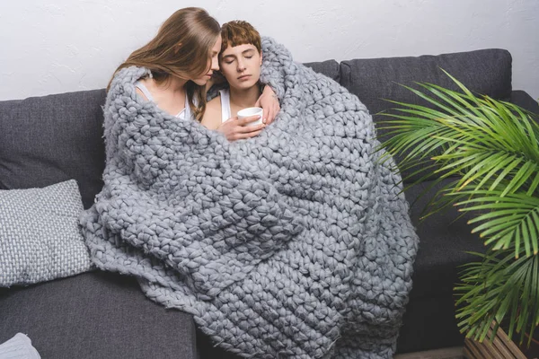 Jovem casal lésbico sentado sob cobertor de lã de malha no sofá com xícara de café — Fotografia de Stock