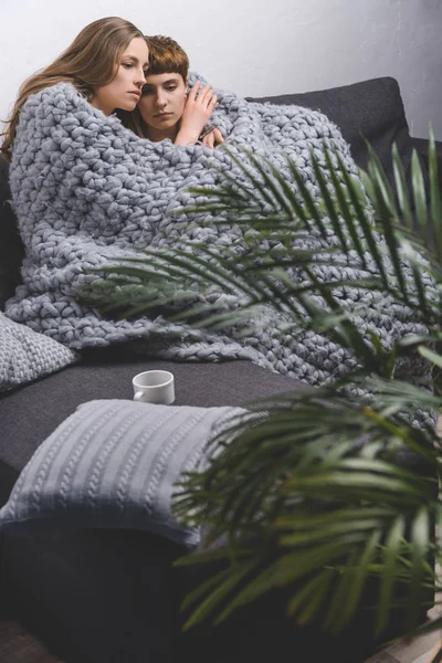 Young lesbian couple sitting under knitted wool blanket together and embracing on couch — Stock Photo
