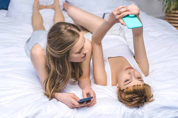 Atractiva pareja de lesbianas jóvenes en red con teléfonos inteligentes en la cama - foto de stock