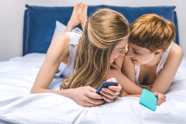 Feliz joven pareja lesbiana abrazos en la cama mientras se utiliza teléfonos inteligentes - foto de stock