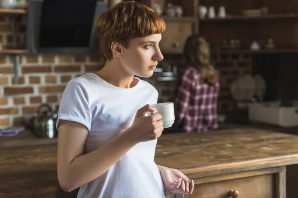 Mujer joven bebiendo café en la cocina y mirando hacia otro lado mientras su novia lava platos - foto de stock