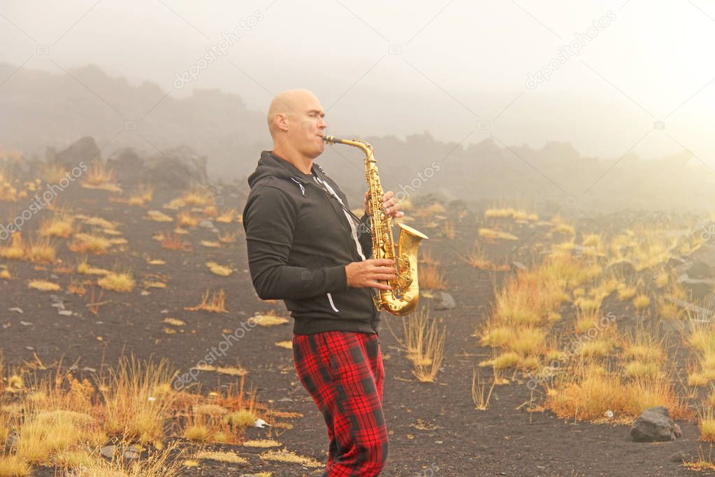 A bald man plays on a gold alto saxophone in the nature, against the backdrop of the volcano Etna, in the fog. Romantic saxophone. Jazz. The man is a musician. The island of Sicily, Italy.