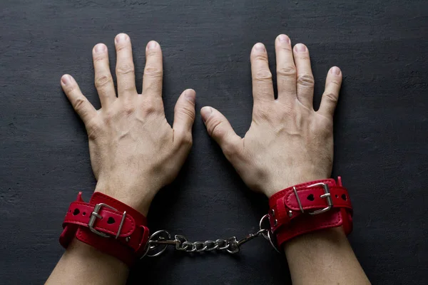 Adult male hands in red erotic leather handcuffs. Hands in captivity. Red leather handcuffs on black background. Adult games and toys. BDSM, fetish wear and kinky sex toy concept with close up.