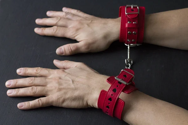 Adult male hands in red erotic leather handcuffs. Hands in captivity. Red leather handcuffs on black background. Adult games and toys. BDSM, fetish wear and kinky sex toy concept with close up.