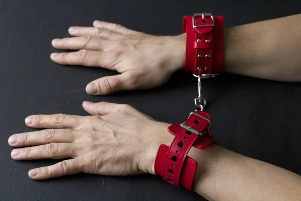 Adult male hands in red erotic leather handcuffs. Hands in captivity. Red leather handcuffs on black background. Adult games and toys. BDSM, fetish wear and kinky sex toy concept with close up.