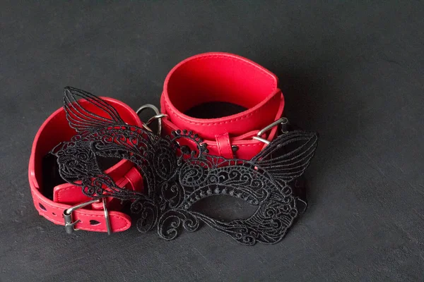 Red leather handcuffs, erotic black mask on the face on black background. Adult games and toys. BDSM, bondage play, fetish wear and kinky sex toy concept with close up.