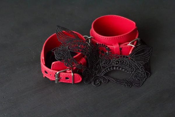 Red leather handcuffs, erotic black mask on the face on black background. Adult games and toys. BDSM, bondage play, fetish wear and kinky sex toy concept with close up.