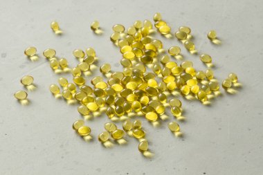 Altın sarısı renkli saydam kapsül yığını açık gri arka planda yatmaktadır. Yağlı kapsüller A vitamini, D3 vitamini, balık yağı, omega 3, 6, 9, akşam çuha çiçeği, D vitamini, E vitamini..