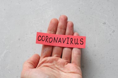 Bir el, açık gri modern arkaplanda kırmızı bir sayfa üzerine yazılmış Coronavirus sözcüğünü tutar. Salgın, tehlike, iyileşme.