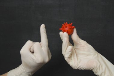 Hand in white medical disposable rubber latex glove shows middle finger to red coronavirus in another rubber glove. Fuck you. Protective disposable gloves protect against viruses, coronavirus.