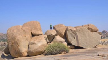 Açık bir yaz gününde, Avrupa görünümlü kel bir adam Hindistan, Hampi 'de çok büyük taşların üzerinde duruyor, siyah pantolon ve yeşil bir tişört..