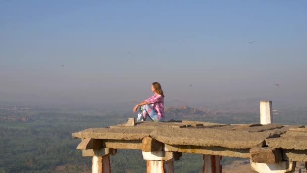 一位身穿紧身衣和粉红格子衬衫的年轻姑娘 长着淡的短发 欧洲风貌 紧闭双眼 坐在一座古印度寺庙的屋顶上 四周都是飞舞的蜻蜓昆虫 — 图库视频影像