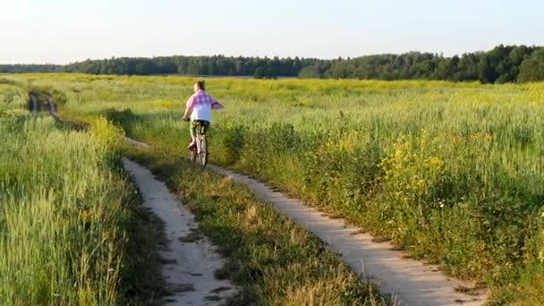 一个年轻的欧洲姑娘骑着自行车穿过夏天的绿地 穿着绿色的裤子 粉色格子衬衫和太阳镜 面带微笑 骑自行车 户外活动 体育娱乐活动 — 图库视频影像