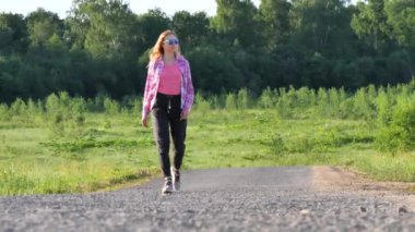 Uzun sarı saçlı, koyu gri spor pantolonlu ve pembe tişörtlü, ince, genç bir kız yazın asfalt bir yolda yürüyor, yüzünü ekrana çeviriyor. Yazın yolda yürü..