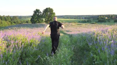 Siyah giysili mutlu genç kel adam, bol tişörtlü pantolon yeşil bir çiçek tarlasında koşuyor ve ayaktan ayağa zıplıyor. Arkadan bak. Özgür bir adam hayattan zevk alır. Yavaş çekim videosu.
