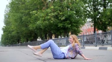 Drunk young girl with long blond red hair in blue trousers, sitting on her ass on the roadway in the city early in the morning, swinging in different directions, raising her legs, smiling. Crazygirl.