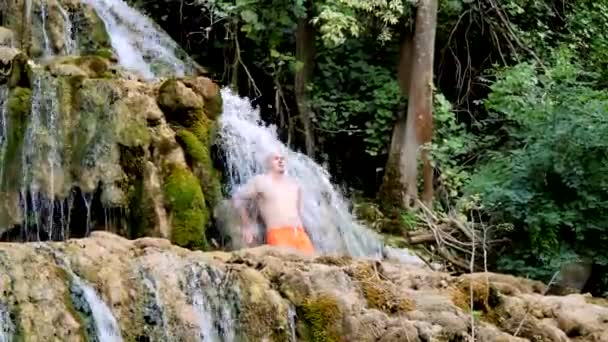 克罗地亚克尔卡国家公园 一个穿着鲜艳橙色短裤的年轻秃头男子站在一个强大的瀑布下 冰水变硬 漂亮的男性身体 健康的生活方式 — 图库视频影像