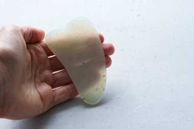 Genç bir kadın el guacha kazıyıcısını tutuyor, yüz masajı için, vücut, doğal beyaz yeşim taşı, beyaz arka plan. Güzellik ve gençlik için terapi masajı. Metnin için boşluk kopyala.