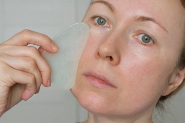 Yüz masajı. Genç bir kız doğal bir mineral için beyaz bir gouacha kazıyıcısı tutuyor ve yakın plan yüz masajı yapıyor. Güzellik ve gençlik için terapi masajı. Metnin için boşluk kopyala.