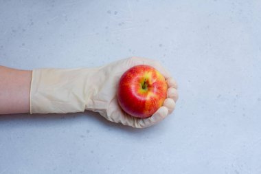 Kırmızı elma, açık gri ve beyaz modern beton arka planda beyaz, tek kullanımlık lastik eldiven içinde duruyor. Lastik eldivendeki bir el elma meyvesi tutar. Coronavirüs salgını konsepti.