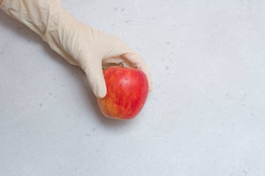 Kırmızı elma, açık gri ve beyaz modern beton arka planda beyaz, tek kullanımlık lastik eldiven içinde duruyor. Lastik eldivendeki bir el elma meyvesi tutar. Coronavirüs salgını konsepti.