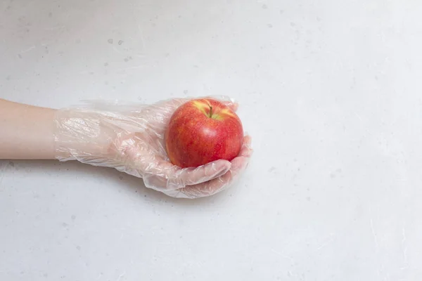 Hand in disposable transparent glove holds red apple fruit. A red apple lies on hand in disposable transparent glove, on light gray and white modern concrete background. Coronavirus epidemic concept.