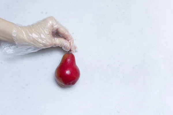 Hand in disposable transparent glove holds Red pear fruit. Red pear lies on hand in disposable transparent glove, on light gray, white modern concrete background. Coronavirus epidemic concept.