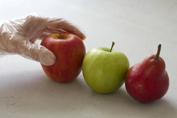 Hand in disposable transparent glove holds touches fruits of red and green apple, red pear, close-up, on a light gray and white modern concrete background. Coronavirus epidemic concept.