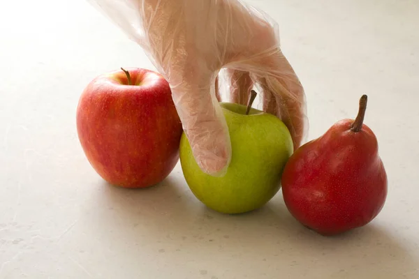 Hand in disposable transparent glove holds touches fruits of red and green apple, red pear, close-up, on a light gray and white modern concrete background. Coronavirus epidemic concept.