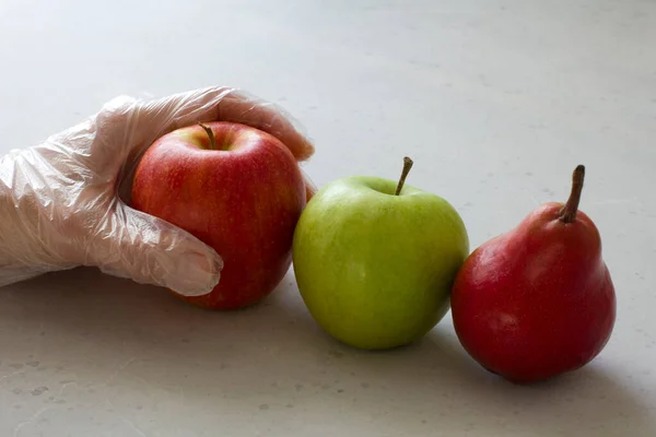 Hand in disposable transparent glove holds touches fruits of red and green apple, red pear, close-up, on a light gray and white modern concrete background. Coronavirus epidemic concept.